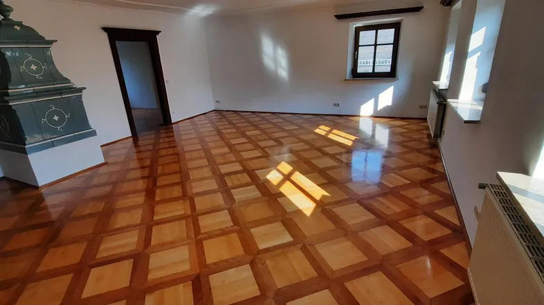 Helles Wohnzimmer mit gemustertem Parkettboden, weiß gestrichenen Wänden und einem traditionellen Kachelofen.