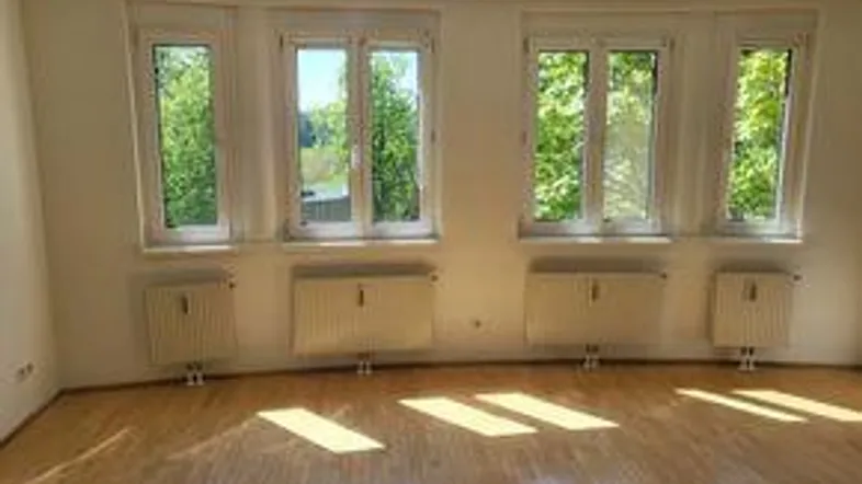 Helles Zimmer mit Parkettboden und mehreren Fenstern, die viel Tageslicht hereinlassen.