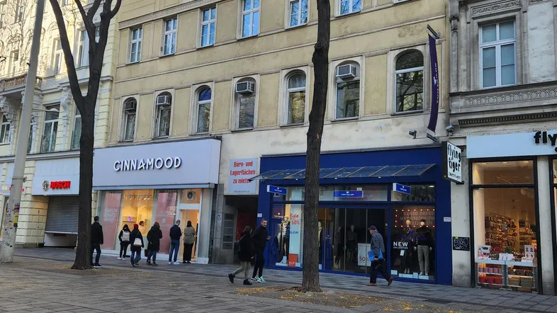 Gebäude an einer belebten Straße mit Geschäften im Erdgeschoss und Wohnungen darüber, umgeben von Bäumen.