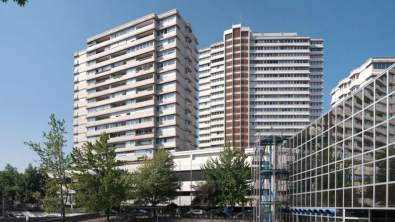 Moderne Hochhäuser mit zahlreichen Balkonen und einer Glasfassade im Vordergrund, umgeben von Bäumen und einem Parkplatz.