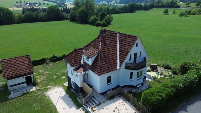 Luftaufnahme eines Hauses mit rotem Ziegeldach und weißer Fassade, umgeben von grünen Feldern und Bergen.
