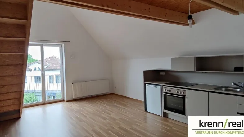 Heller Wohnbereich in einem Dachgeschoss-Apartment mit Holzbalken, Parkettboden und Zugang zu einem kleinen Balkon.