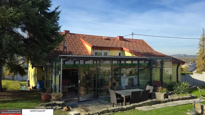 Haus mit Wintergarten, gepflasterter Terrasse und Gartenmöbeln unter blauem Himmel.