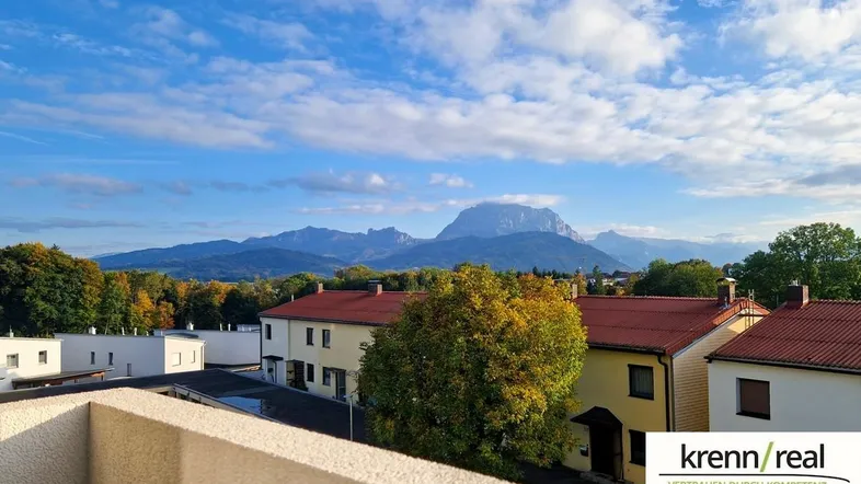 Weitblick über Dächer und herbstliche Bäume mit majestätischem Bergpanorama unter blauem Himmel.
