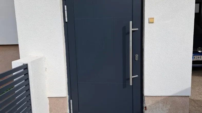 Moderne dunkelgraue Eingangstür mit Edelstahlgriff und weißer Putzfassade, davor ein gepflasterter Weg.