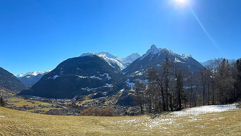Weitläufiger Panoramablick auf schneebedeckte Berge und ein grünes Tal unter strahlend blauem Himmel.