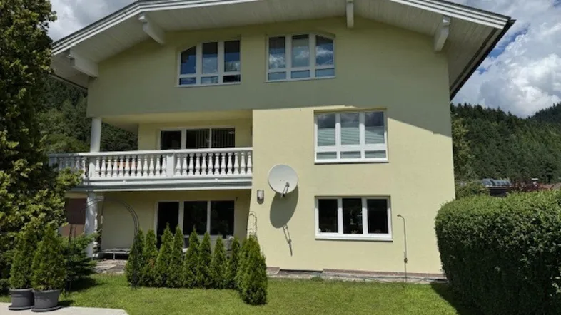 Hausfassade in hellem Gelb mit Balkon und gepflegtem Vorgarten unter bewölktem Himmel.