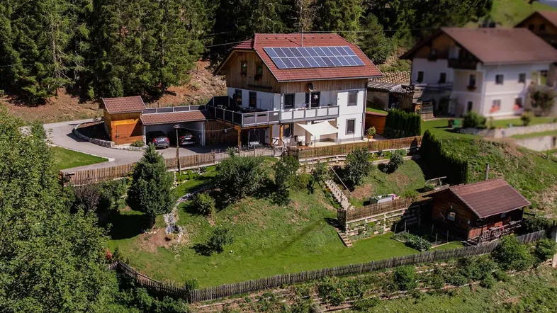 Luftaufnahme eines Hauses mit Solaranlage, großer Terrasse und gepflegtem Garten in bergiger Umgebung.