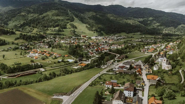 Panoramablick aus der Vogelperspektive auf ein malerisches Dorf, umgeben von grünen Hügeln und bewaldeten Bergen.