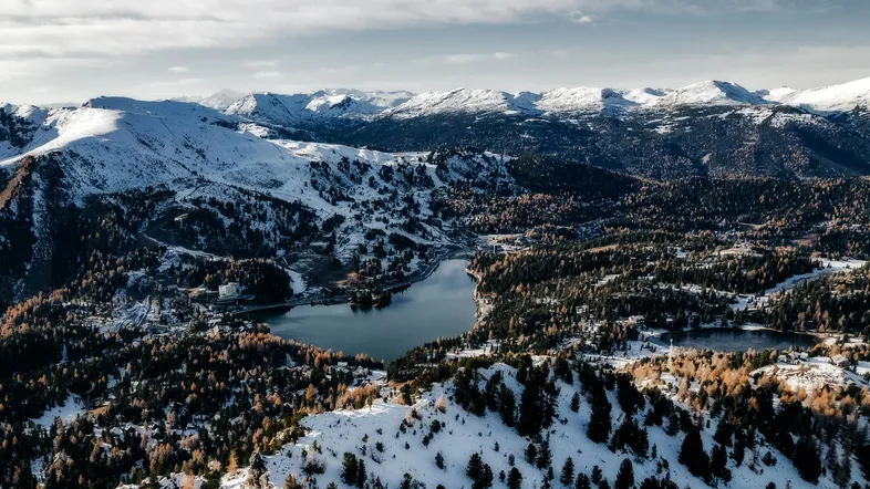 Panoramablick aus der Vogelperspektive auf schneebedeckte Berge, einen See und dichte Wälder.