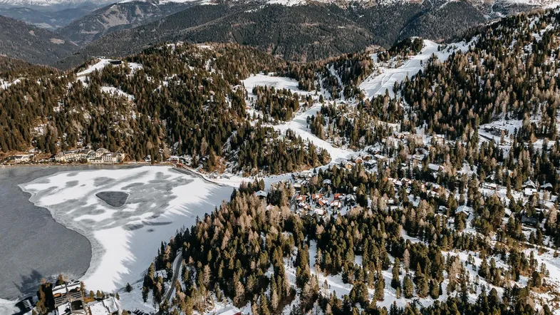 Luftaufnahme einer verschneiten Berglandschaft mit zugefrorenem See, Wäldern und Skigebiet.