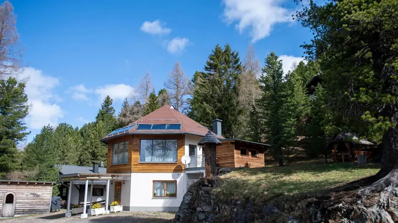 Einzigartiges achteckiges Haus mit Holzfassade und großen Fenstern, umgeben von Nadelbäumen unter blauem Himmel.