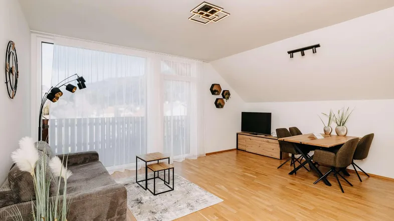Helles Wohn- und Esszimmer mit Holzboden, gemütlichem Sofa und Zugang zum Balkon mit Bergblick.