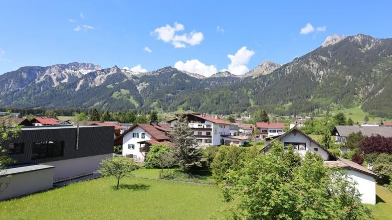 Weitläufiger Blick über die grüne Landschaft und das Dorf mit majestätischen Bergen im Hintergrund.