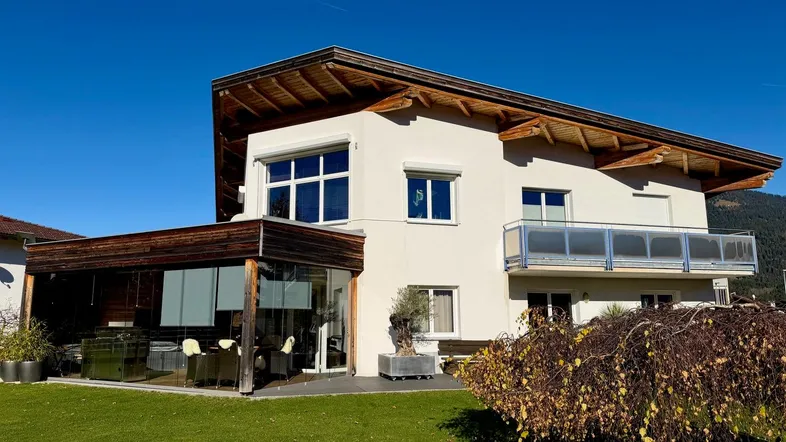 Modernes Einfamilienhaus mit großem Garten, verglaster Terrasse und Balkon unter blauem Himmel.