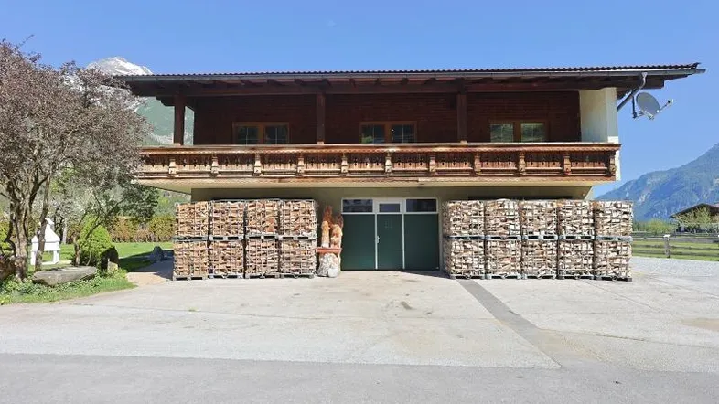 Traditionelles Holzhaus mit Balkon und gestapeltem Brennholz im Vordergrund, umgeben von Bergen.