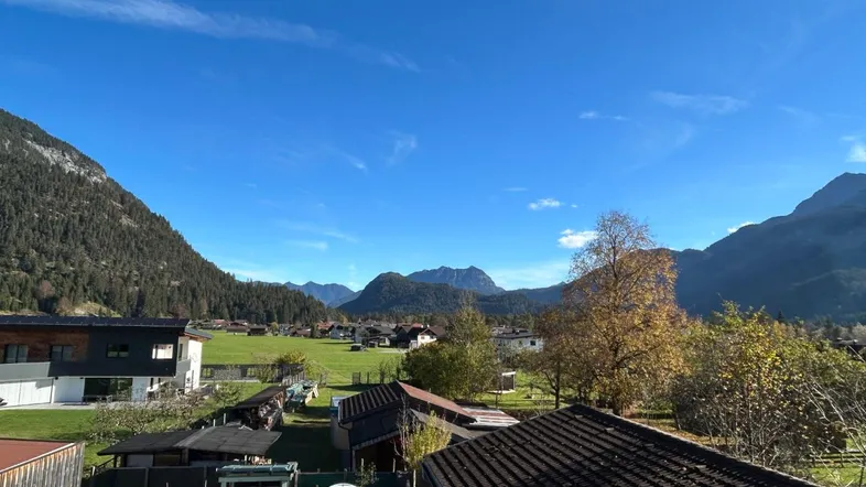 Weitläufiger Blick über grüne Felder und Dächer auf die umliegende Berglandschaft unter blauem Himmel.