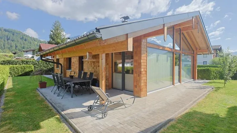 Moderne Holzhausfassade mit großer gepflasterter Terrasse und Gartenmöbeln unter blauem Himmel.