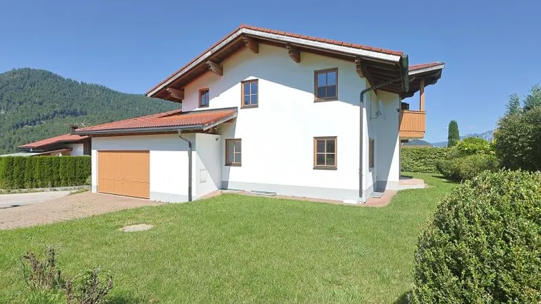 Freistehendes Einfamilienhaus mit weißer Fassade, braunem Dach und integrierter Garage, umgeben von Grünflächen und Bergen.