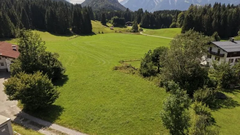 Weitläufiges grünes Feld mit majestätischen Bergen und dichten Wäldern im Hintergrund, umgeben von ländlicher Bebauung.