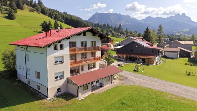 Großes Wohnhaus mit rotem Satteldach und Balkonen, umgeben von grünen Wiesen und Bergen.