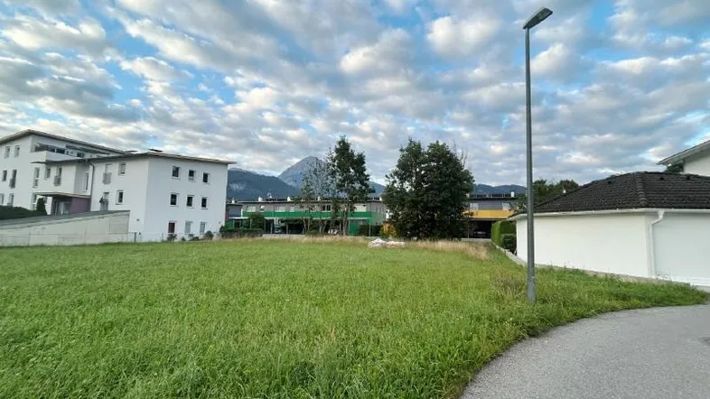 Weitläufiges Grundstück mit grüner Wiese, umgeben von Gebäuden und mit Blick auf die Berge unter bewölktem Himmel.