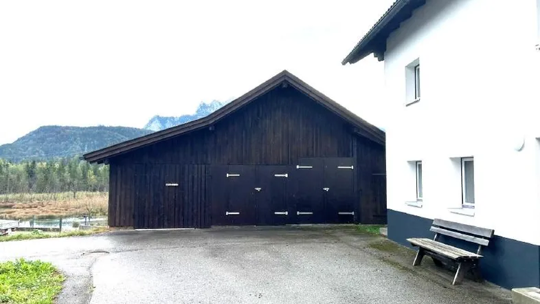 Außenansicht eines weißen Gebäudes neben einer dunklen Holzscheune mit Bergblick und Teich im Hintergrund.
