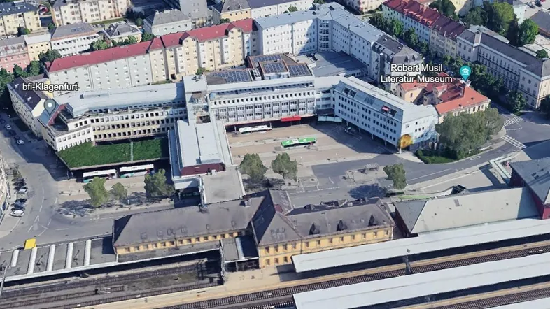 Luftaufnahme eines Stadtviertels mit mehreren Gebäudekomplexen, Straßen und Bahngleisen, umgeben von Bäumen.