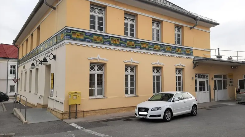 Gelbes Gebäude mit traditionellen Fenstern und dekorativem Fries, davor ein geparktes Auto und eine Rampe.