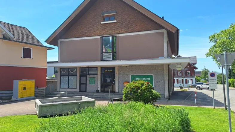 Gebäudefront einer Tierarztpraxis mit Giebeldach, großen Fenstern und Grünfläche unter blauem Himmel.