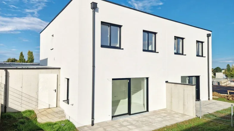 Modernes Einfamilienhaus mit weißer Fassade, Terrasse und kleinem Gartenbereich unter blauem Himmel.