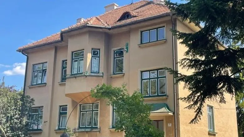 Historische Villa mit beiger Fassade, grünen Fensterrahmen und rotem Ziegeldach unter blauem Himmel.