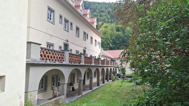 Langes Gebäude mit Arkaden und Balkonen, umgeben von einem grünen Gartenbereich und Bäumen.