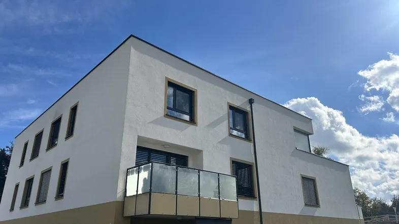 Modernes Gebäude mit weißer und beiger Fassade, schwarzen Fenstern und einem kleinen Balkon unter blauem Himmel.