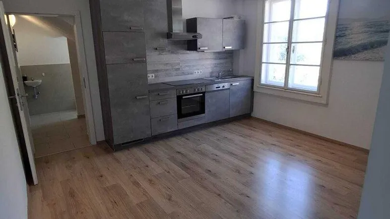 Moderne Einbauküche in Grau mit Holzboden und Fenster, offen zu einem weiteren Raum.