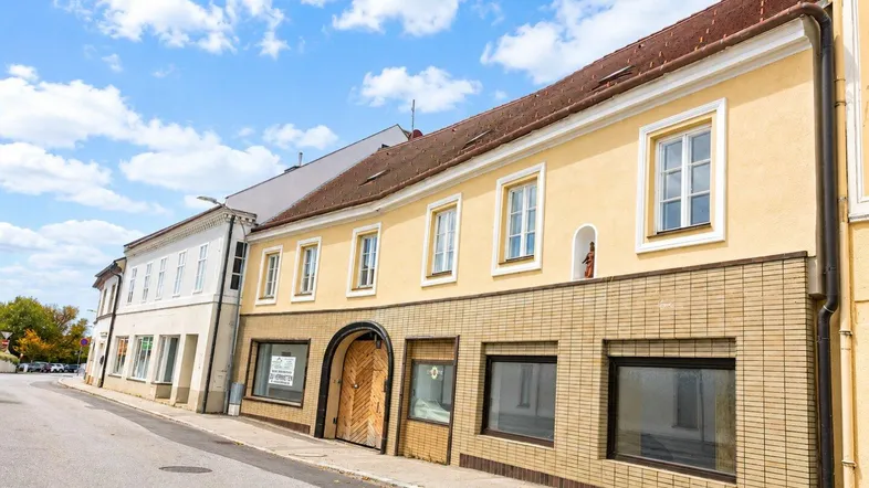Gelbe Fassade eines historischen Gebäudes an einer Straße mit braunen Ziegeln und Holztür unter blauem Himmel.