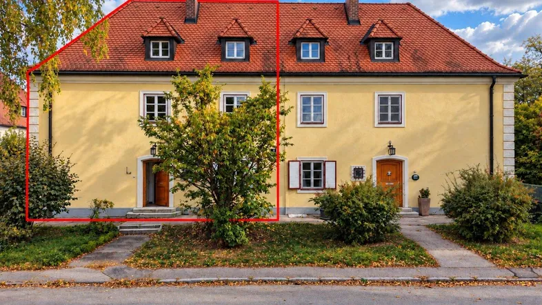 Gelbes Gebäude mit rotem Ziegeldach und mehreren Gaubenfenstern, umgeben von Bäumen und Vorgarten.