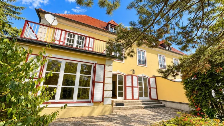 Historisches Gebäude mit gelber Fassade, roten Fensterläden und einem Balkon im Obergeschoss.