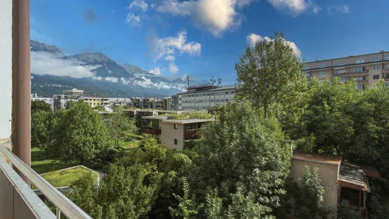 Panoramablick vom Balkon auf die umliegende Stadtlandschaft mit grünen Bäumen und majestätischen Bergen im Hintergrund.