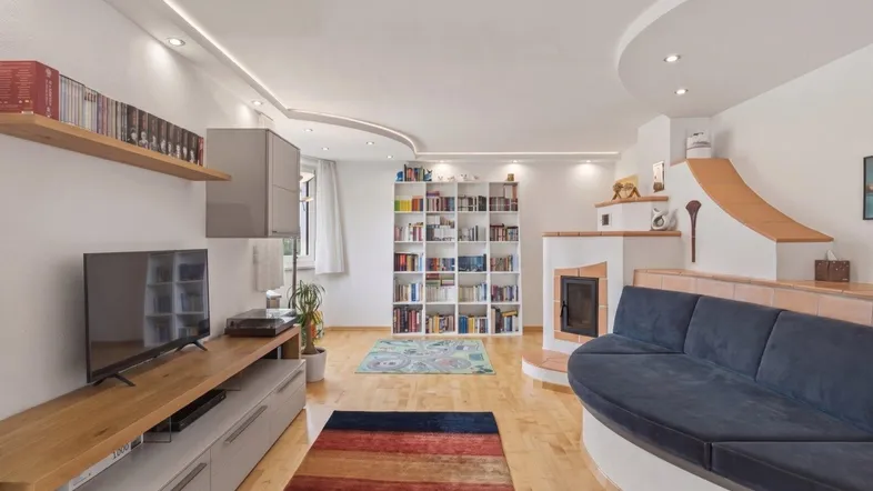 Gemütliches Wohnzimmer mit integriertem Sofa, Kachelofen, Bücherregalen und hellem Parkettboden.