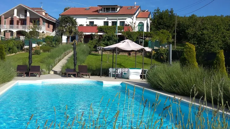 Großer Außenpool mit Liegestühlen und einem Pavillon, umgeben von grüner Landschaft und Häusern.