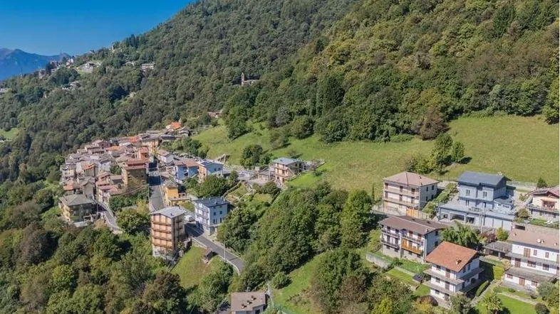 Luftaufnahme eines malerischen Dorfes am Berghang, umgeben von üppiger grüner Natur und weiteren Häusern.