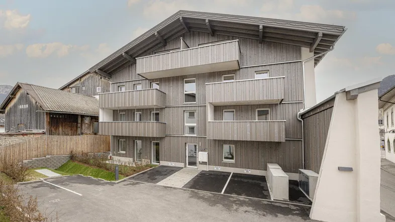 Modernes Mehrfamilienhaus mit Holzfassade, Balkonen und Außenstellplätzen in alpiner Umgebung.