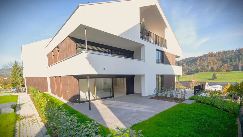 Modernes Neubau-Einfamilienhaus mit weißer Fassade, Holzdetails und großzügiger Terrasse, umgeben von Grünflächen.