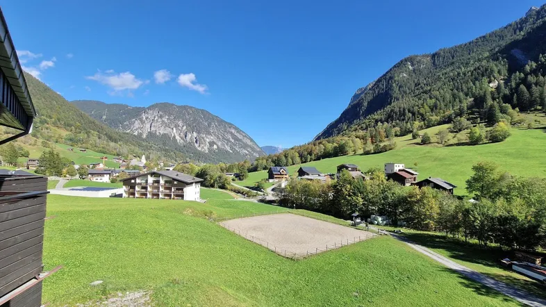 Weitläufiger Blick über ein grünes Tal mit Bergen, Häusern und einem Reitplatz unter blauem Himmel.