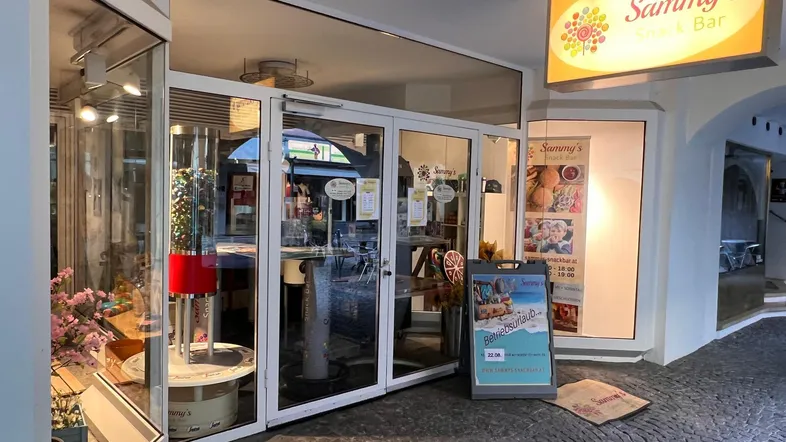 Eingangsbereich einer Snackbar mit großen Glasfenstern und Leuchtreklame, die den Namen des Geschäfts zeigt.