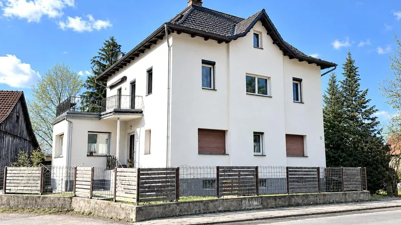 Weißes Einfamilienhaus mit Satteldach, kleinem Balkon und gepflegtem Vorgarten.