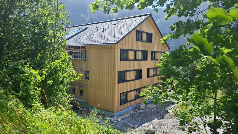 Neubau mit Holzfassade und dunklem Dach, umgeben von grüner Natur und Bergen.