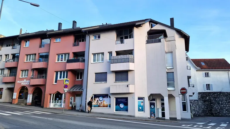 Mehrere Gebäude entlang einer Straße mit Geschäften im Erdgeschoss und Wohnungen darüber, unter einem klaren Himmel.