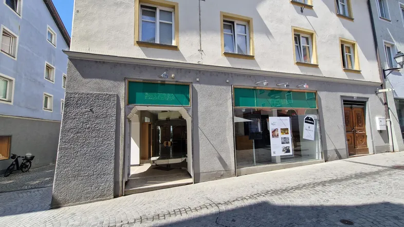 Vorderansicht eines Geschäftsgebäudes an einer gepflasterten Straße mit großen Schaufenstern und Eingang.